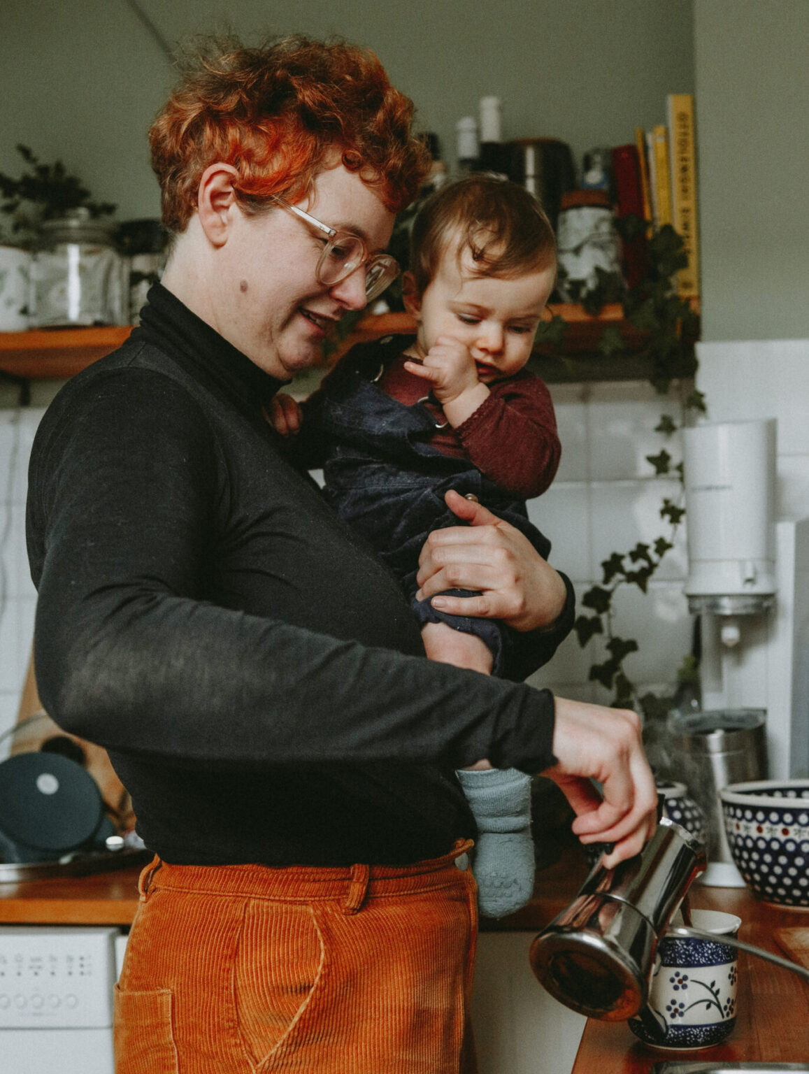 Mutter, die mit Tochter auf dem Arm Kaffee zubereitet.